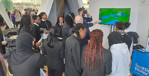 Pupils wearing black school uniform gather round a Scottish Water employee wearing a VR headset. 