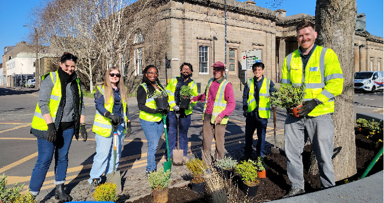 A group of seven people wearing high-visibility vests and gloves are planting greenery around a tree in an urban setting. They are standing on a paved area with a mix of potted plants, soil, and gardening tools. The background features a historic stone building with large windows and columns, as well as a clear blue sky. 
