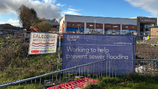 A construction site fenced off with Scottish Water signage reading “Working to help prevent sewer flooding.” Another banner for the Caledonia Water Alliance hangs on the fence. Behind the site is a retail building with signs for outdoor and sports shops. The sky is partly cloudy with trees visible to the left.