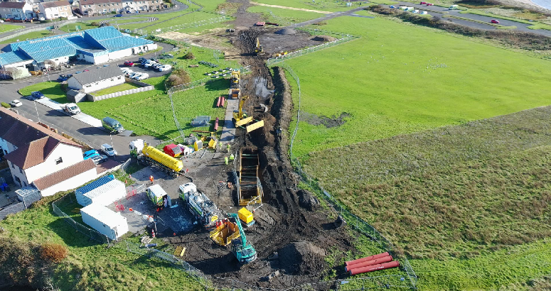 Aerial view of a construction site where repair work is underway on a large underground pipe. Excavators, machinery, and work vehicles are positioned along a long trench through a grassy area near houses. Workers in high-visibility clothing are on site, with safety barriers and temporary equipment set up around the work zone.
