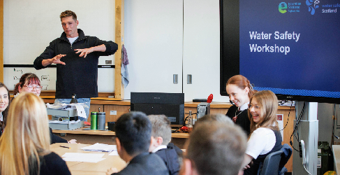 Duncan Scott stands at the front of a classroom presenting. Pupils sit smiling and laughing in the foreground.