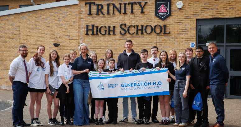 Group photo of students and staff standing outside Trinity High School holding a banner that reads 'Scottish Water - We won the Generation H₂O Making Waves Challenge'. Everyone is smiling and facing the camera.