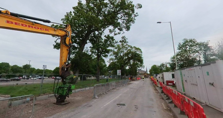 A grey road runs down the centre of the image. To the right is orange plastic fencing, and to the left is a yellow piece of heavy plant machinery with trees in the background.