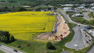 Aerial view of a vibrant landscape showing a large bright yellow rapeseed field alongside roads with moving vehicles and construction equipment near a developing area. Rolling hills and greenery are visible in the background under a clear sky.