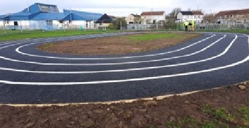 Tarmac of a running track with lines is in the foreground. Houses can be seen in the background with a cloudy blue sky.