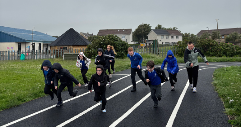 Children running along a running track. Grass surrounds the track and buildings can be seen in the background. The sky is grey.