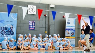 Group of children in swim caps seated on the poolside during a swimming class at a public aquatic facility, with instructors standing by and promotional banners visible in the background.