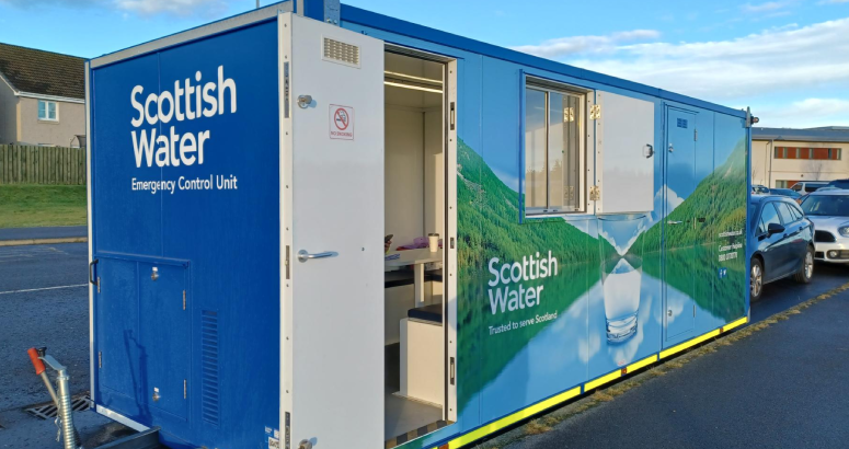 A mobile customer unit branded with Scottish Water logo and with an open white door. It's sat outside on a grey road with pavement in foreground and blue skies in background.