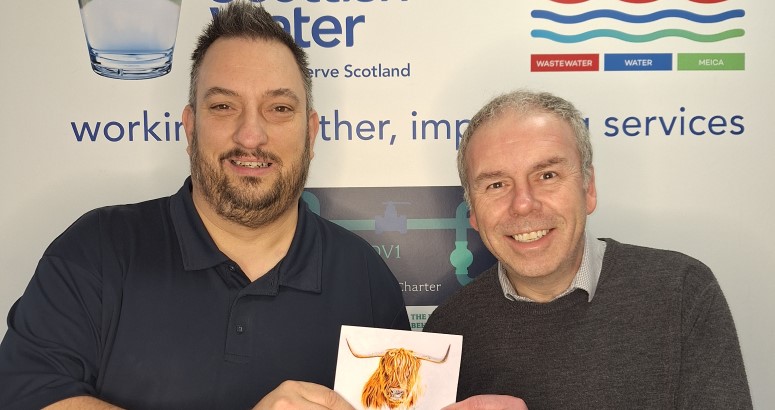 Two individuals standing in front of a Scottish Water banner, one holding a Thank You card from a customer with an image of a highland cow on it.