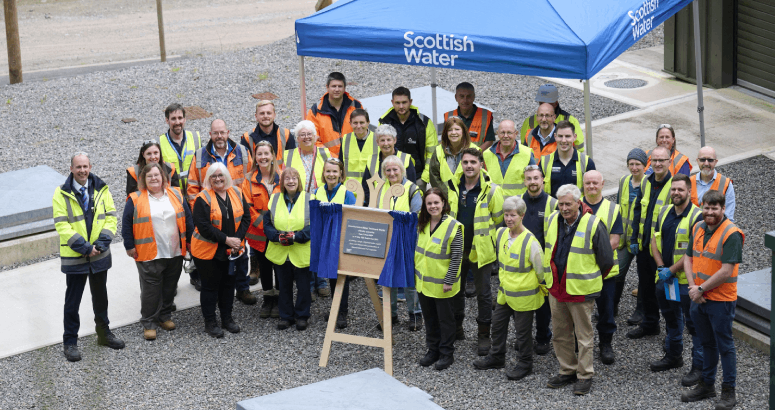 A large group of people wearing hi-viz PPE are standing outdoors, behind a wooden easel with stone plaque.