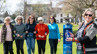 A group of people stand with their arms linked, facing towards the camera while standing on grassed area. On the right hand side, a person stands holding a water bottle and fills it under a blue Scottish Water top up tap.