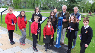 A group of children and adults stand outside. They are holding blue water bottles and surrounding a blue Scottish Water outdoor tap.