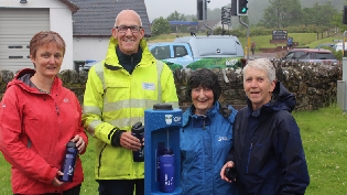 A group of people are standing outside surrounding a blue Scottish Water branded water tap. They are holding blue plastic bottles and facing the camera. In the background is a white building with grey roof, and a Scottish Water branded vehicle.