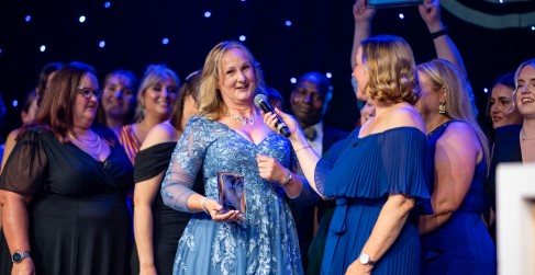 Recipient in a blue dress smiling and holding an award at a ceremony, surrounded by applauding attendees on a stage with a starry backdrop.