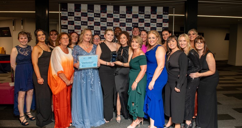 Group of individuals holding an award at a formal event, posing in front of a banner with logos.