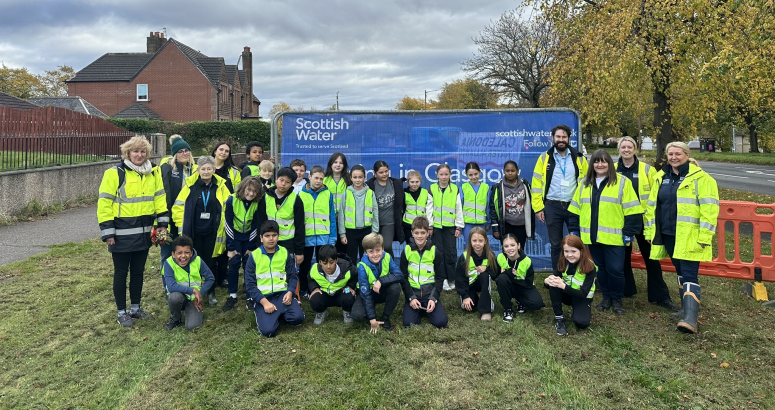 Group of people of various ages wearing safety vests, standing in front of a Scottish Water truck, in a suburban area.