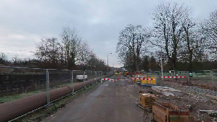 Construction site on a partially closed Shore Road with barriers and fencing; pipes and construction materials are visible along the road, with trees and overcast skies in the background.