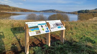 2 new signs side by side at Carron Valley on a sunny day with reservoir in background.
